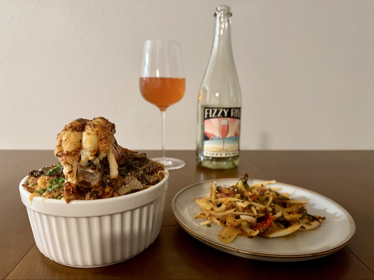 Baked lobster mac and cheese in a ramekin topped with a lobster tail, next to a pepper salad plate and a bottle of sparkling wine